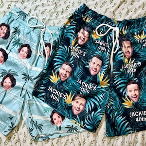 May include: Two pairs of swim shorts with drawstring waistbands. One pair is light blue with a palm tree pattern and repeating faces. The other pair is dark green with a tropical leaf pattern, repeating faces, and the text Jackie's 40th.