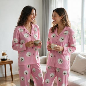 May include: A pink long-sleeved pyjama set featuring a repeating pattern of white dog faces. The top has a collared neckline with a button-down front, and the matching trousers have an elasticated waistband. The fabric is a soft, lightweight material.