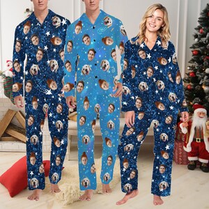 Op de afbeelding: Drie mensen dragen blauwe pyjama's met een wit sterrenpatroon en verschillende gezichten erop gedrukt. De pyjama's hebben een knoopsluiting aan de voorkant en lange mouwen. De mensen staan voor een kerstboom en een open haard.