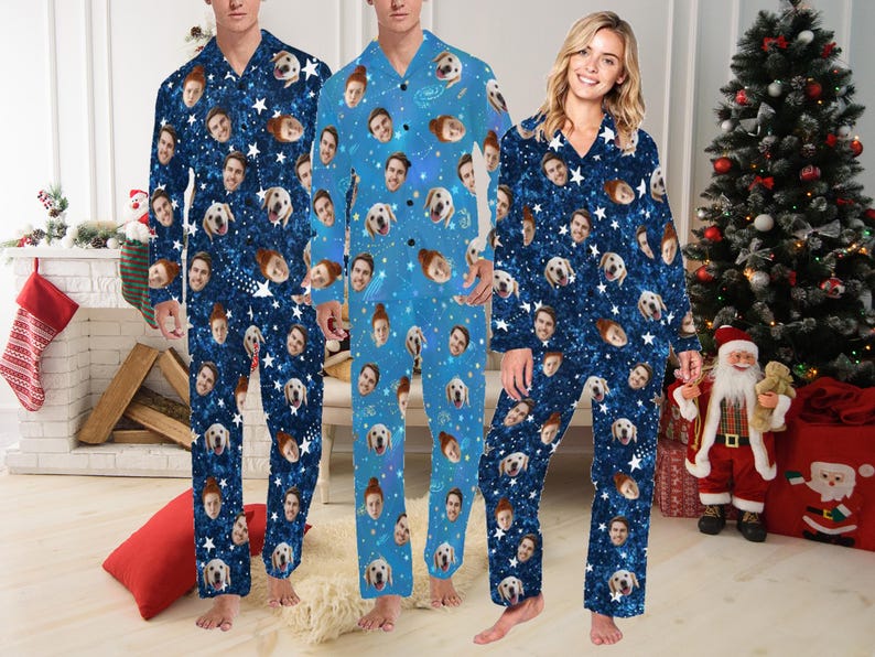 Op de afbeelding: Drie mensen dragen blauwe pyjama's met een wit sterrenpatroon en verschillende gezichten erop gedrukt. De pyjama's hebben een knoopsluiting aan de voorkant en lange mouwen. De mensen staan voor een kerstboom en een open haard.