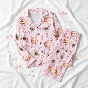 May include: A light pink pyjama set consisting of a long-sleeved button-down shirt and matching trousers. The fabric features a repeating pattern of smiling baby faces wearing headbands and white dogs, interspersed with pink martini glass icons.