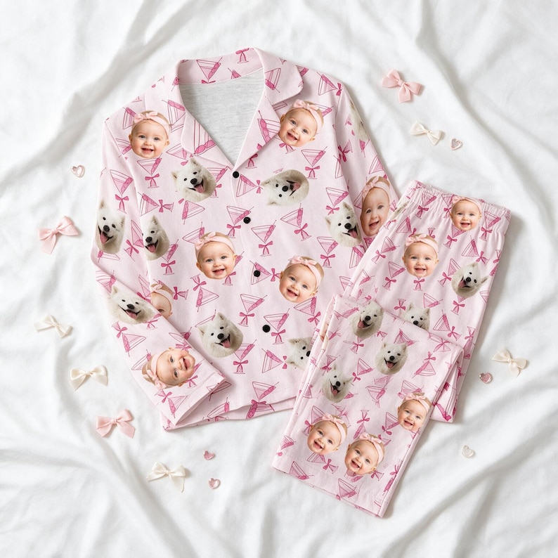 May include: A light pink pyjama set consisting of a long-sleeved button-down shirt and matching trousers. The fabric features a repeating pattern of smiling baby faces wearing headbands and white dogs, interspersed with pink martini glass icons.