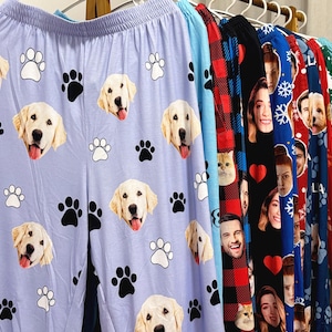 May include: A variety of pajama pants hanging on a rack. One pair is light blue with a pattern of golden retriever faces and paw prints. Other pairs feature faces, hearts, and plaid patterns.
