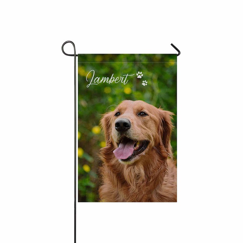 May include: A garden flag displaying a Golden Retriever with its tongue out. The flag has the name "Jamber" in script with paw prints. The background is a blurred green with yellow flowers, and the flag is attached to a black metal pole.