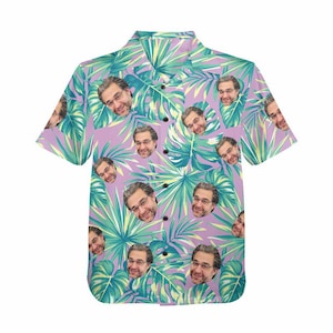 May include: A short-sleeve button-down shirt featuring a repeating pattern of a man's face with glasses and a beard, interspersed with large green and light green tropical palm leaves against a solid light purple background.