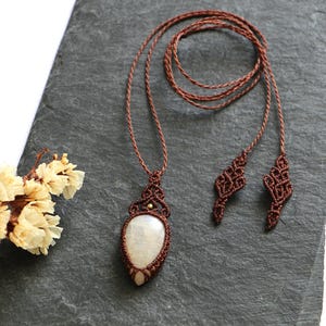 May include: A handcrafted macramé necklace with a large, oval, off-white stone pendant. The necklace features intricate knotting in a brown cord, with decorative elements at the ends. A small bouquet of dried flowers rests near the necklace on a dark grey surface.