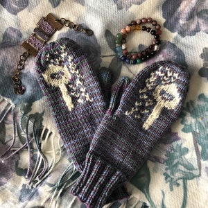 Peut inclure: Une paire de mitaines en tricot violet et blanc avec un motif d'arbre. Les mitaines sont posées sur un tissu à motifs avec un bracelet marron et un bracelet en perles.