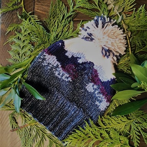 May include: A blue, white and purple knitted hat with a white pom-pom. The hat is surrounded by green foliage.