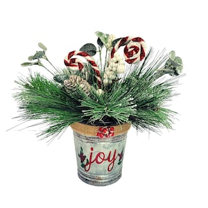 May include: A festive Christmas centerpiece featuring a galvanized metal bucket with the word "joy" painted on it. The bucket is filled with green pine branches, white berries, and red and white candy canes.