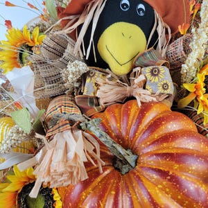 Puede incluir: Una corona de puerta con tem&aacute;tica oto&ntilde;al con un espantap&aacute;jaros negro y amarillo, una calabaza naranja grande y girasoles.