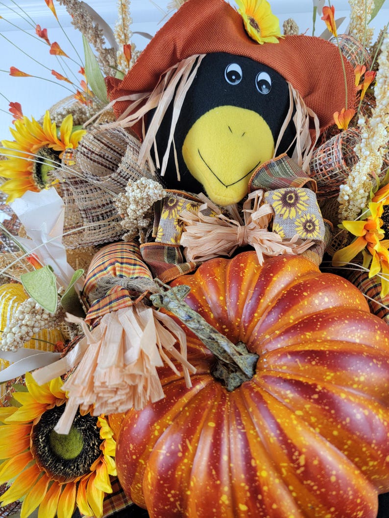 Puede incluir: Una corona de puerta con tem&aacute;tica oto&ntilde;al con un espantap&aacute;jaros negro y amarillo, una calabaza naranja grande y girasoles.