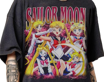 May include: Dark grey t-shirt with a vibrant graphic of Sailor Moon characters. The design includes the text "SAILOR MOON" in a retro style. The artwork features multiple anime figures with yellow hair, sailor outfits, and dynamic poses.