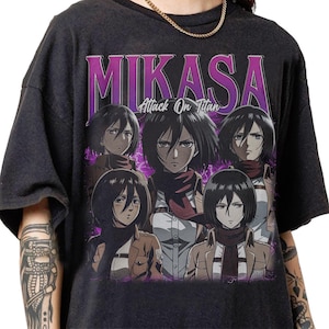 May include: Dark gray t-shirt featuring a graphic of Mikasa from Attack on Titan. The design includes multiple images of the character with the name "MIKASA" in large, purple letters above the images. The text "Attack On Titan" is below the name.