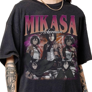 Könnte beinhalten: Dunkelgraues T-Shirt mit einer Grafik von Mikasa Ackerman aus der Anime-Serie Attack on Titan. Das Design zeigt den Namen "MIKASA" in einer Retro-Schriftart, darunter "Ackerman". Das Bild zeigt Mikasa in verschiedenen Posen mit einem lila-orangenen Hintergrund.