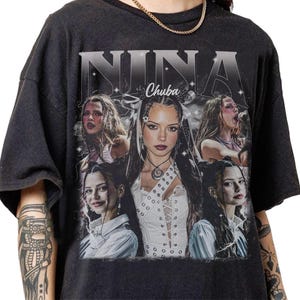 May include: Dark gray t-shirt featuring a collage of images of a woman, with the name "NINA" in large, stylized letters above the images. The word "Chuba" is below the name. The woman is wearing a white top in the central image.