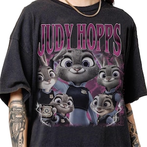 May include: Dark gray t-shirt featuring a graphic of Judy Hopps from Zootopia. The design includes the name "JUDY HOPPS" in a pink, stylized font above an image of Judy Hopps and other characters from the movie.