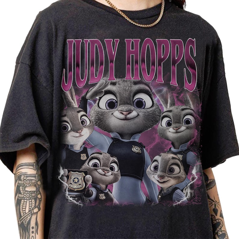 May include: Dark gray t-shirt featuring a graphic of Judy Hopps from Zootopia. The design includes the name "JUDY HOPPS" in a pink, stylized font above an image of Judy Hopps and other characters from the movie.
