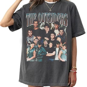 May include: A charcoal gray short-sleeve t-shirt featuring a graphic print of the cast of the movie The Outsiders. The design includes the text The Outsiders in a stylized font above a collage of the actors with a lightning bolt background.