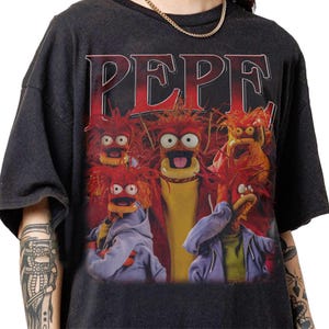 May include: Dark gray t-shirt featuring a graphic of the Muppet character Pepe the King Prawn and other characters. The word "PEPE" is printed in red across the top. The characters have red hair and are wearing various outfits.