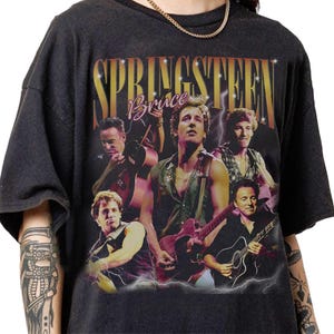 May include: Dark gray t-shirt featuring a multi-image design of Bruce Springsteen with guitars and the name "SPRINGSTEEN" in gold lettering. The word "Bruce" is in a cursive font. Lightning bolt graphics are also present.