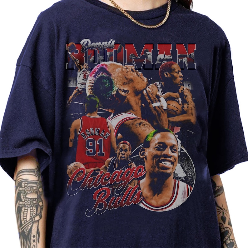 May include: Navy blue t-shirt featuring a collage of images of Dennis Rodman, including action shots and his name. The shirt also displays the words "Chicago Bulls" in a cursive font, and Rodman's jersey number, 91.