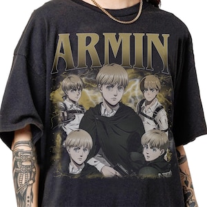 May include: Dark gray t-shirt featuring the name "ARMIN" in gold lettering above a graphic of a character from an anime series. The graphic shows the character in multiple poses against a background of lightning.