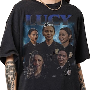 May include: Black t-shirt featuring a graphic of a woman with the name "LUCY Chen" in blue text. The graphic includes multiple images of the woman in a police uniform against a backdrop of lightning.