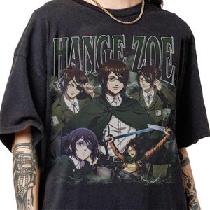 Pode incluir: Camiseta cinza escuro com um gráfico de personagens de anime e o texto "Hange Zoe" em verde. O design inclui várias figuras em uniformes militares, com uma segurando uma espada. A camiseta tem um visual vintage e desgastado.