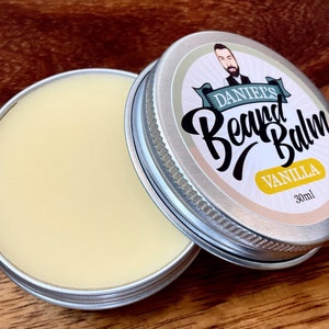 May include: A silver tin with a white label that says "Daniel's Beard Balm Vanilla 30ml". The label has a cartoon illustration of a man with a beard. The tin is open and the balm is a light yellow color.