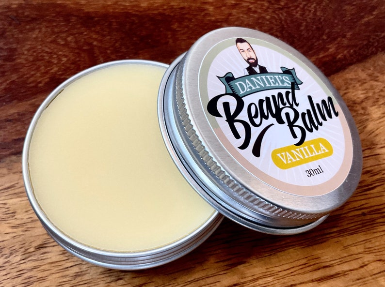 May include: A silver tin with a white label that says "Daniel's Beard Balm Vanilla 30ml". The label has a cartoon illustration of a man with a beard. The tin is open and the balm is a light yellow color.