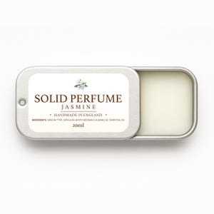 Op de afbeelding: Een zilveren rechthoekige doos met vaste jasmijnparfum, gedeeltelijk open. Het etiket luidt "SOLID PERFUME JASMINE" met "HANDMADE IN ENGLAND" en ingrediënten. De doos bevat 20 ml parfum.