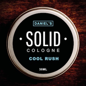Puede incluir: Una lata redonda de Daniel's Solid Cologne, de color plateado. La etiqueta negra presenta el texto blanco "SOLID COLOGNE COOL RUSH" y "30ML". El nombre de la marca está en un recuadro azul. Sobre una superficie de madera oscura.