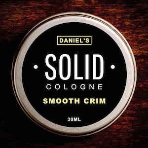 May include: A circular tin of Daniel's Solid Cologne with a silver rim. The black label displays white text: "SOLID COLOGNE SMOOTH CRIM" and "30ml". The tin is set against a dark wooden background.