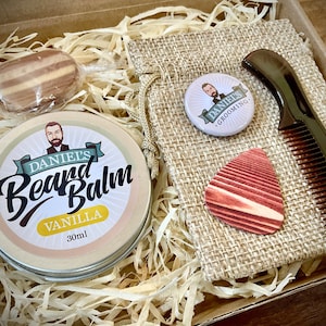 Peut inclure: Une boîte-cadeau remplie de produits de soin de la barbe, dont une boîte de baume à barbe étiquetée "Daniel's Beard Balm Vanilla 30ml", un petit sac en toile de jute avec un badge bouton qui dit "Daniel's Grooming", un médiator en bois et un peigne brun.