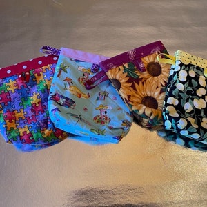 May include: Four fabric drawstring bags with different patterns. The bags are lined up in a row on a silver background. The first bag has a red and white polka dot trim and a colorful puzzle pattern. The second bag has a light blue background with a cartoon pattern of people and animals. The third bag has a dark red background with a sunflower pattern. The fourth bag has a black background with a white and green lemon pattern.