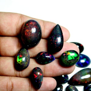May include: A collection of polished black opal gemstones in various shapes, including oval and teardrop, featuring vibrant flashes of red, green, and blue color play against a dark, opaque background, displayed in a group of loose, smooth cabochons.