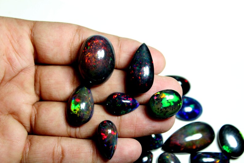 May include: A collection of polished black opal gemstones in various shapes, including oval and teardrop, featuring vibrant flashes of red, green, and blue color play against a dark, opaque background, displayed in a group of loose, smooth cabochons.