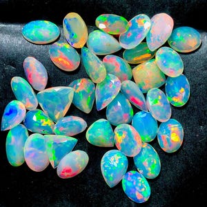 Può includere: Una collezione di opali sfaccettati sciolti in varie forme, inclusi tagli ovali, a goccia e triangolari. Ogni gemma presenta una base traslucida e lattiginosa con vivaci bagliori multicolori di luce blu, verde, arancione e rosa in tutta la superficie.