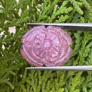 Puede incluir: Una piedra preciosa rosa translúcida tallada en forma de flor con pétalos redondeados y un detalle circular central. La piedra está sujeta por unas pinzas de metal plateado, mostrando su superficie pulida y su diseño en relieve simétrico.