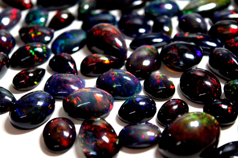 May include: A collection of polished black opal gemstones with smooth, rounded surfaces. Each stone features a dark base color with vibrant, iridescent flashes of red, green, blue, and orange light reflecting from within the cabochon-cut gems.