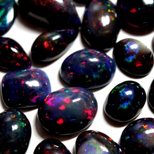 May include: A collection of polished black opals featuring smooth, rounded surfaces and vibrant, multi-colored flashes of red, green, and blue light. These gemstones exhibit a dark, opaque base color with internal iridescent patterns.
