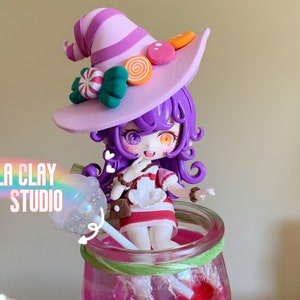 Puede incluir: Una caprichosa figura de arcilla de una niña con cabello morado y un sombrero de bruja adornado con caramelos. La figura está dentro de un frasco de vidrio con una piruleta. El texto "NEBULA CLAY STUDIO" es visible. La figura lleva un vestido a rayas rojas y blancas.