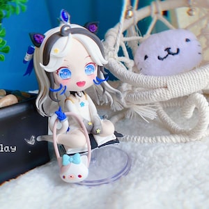 Può includere: Una bambola di stile anime bianca e grigia con occhi azzurri e un fiocco blu nei capelli. Indossa un vestito bianco con stelle blu e bianche. La bambola tiene una piccola borsa rosa e blu con un fiocco. La bambola è seduta su un supporto di plastica trasparente. La bambola è davanti a un piccolo albero in vaso e una sedia a dondolo in vimini bianca.