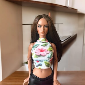 May include: A fashion doll with long brown hair and blue eyes, wearing a white halter top with a floral pattern of pink and blue flowers and green leaves. The doll is also wearing black leather pants.