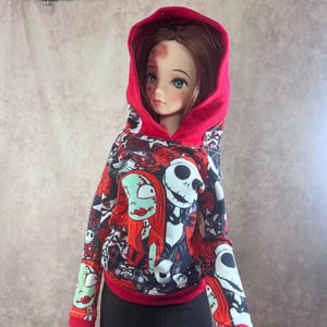 Puede incluir: Una mu&ntilde;eca con una sudadera con capucha roja con un estampado en blanco, negro y rojo con personajes de Pesadilla antes de Navidad. La mu&ntilde;eca tambi&eacute;n lleva pantalones negros.