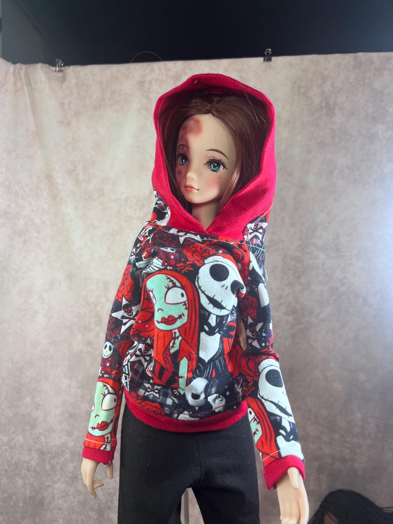 Puede incluir: Una mu&ntilde;eca con una sudadera con capucha roja con un estampado en blanco, negro y rojo con personajes de Pesadilla antes de Navidad. La mu&ntilde;eca tambi&eacute;n lleva pantalones negros.