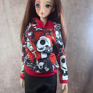Puede incluir: Una mu&ntilde;eca con una sudadera con capucha roja, negra y blanca con un dise&ntilde;o de Jack Skellington y pantalones negros. La mu&ntilde;eca tiene el pelo largo casta&ntilde;o y ojos azules. La sudadera tiene capucha y pu&ntilde;os rojos.