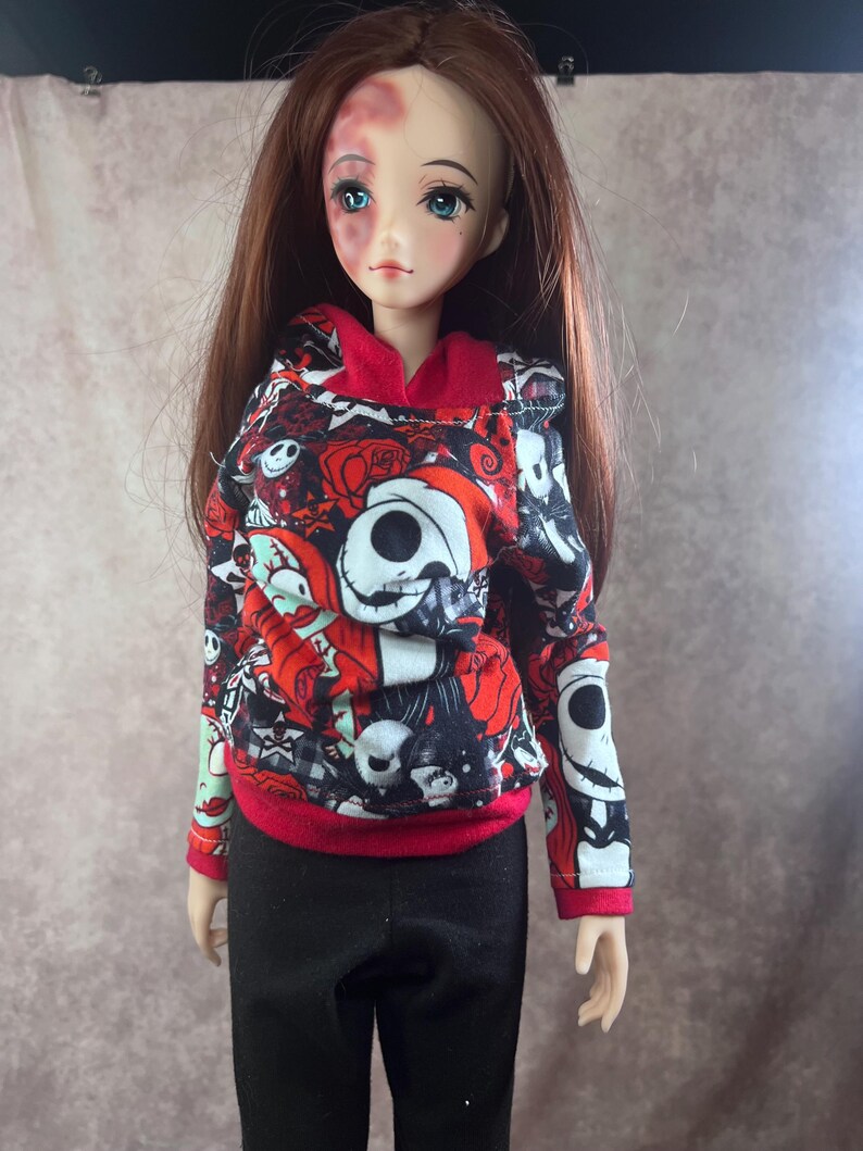 Puede incluir: Una mu&ntilde;eca con una sudadera con capucha roja, negra y blanca con un dise&ntilde;o de Jack Skellington y pantalones negros. La mu&ntilde;eca tiene el pelo largo casta&ntilde;o y ojos azules. La sudadera tiene capucha y pu&ntilde;os rojos.