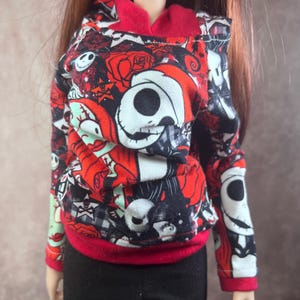 Puede incluir: Una mu&ntilde;eca con una sudadera con capucha estampada en rojo, negro y blanco con capucha y pu&ntilde;os rojos. La sudadera presenta un dise&ntilde;o repetido de calaveras, rosas y otros elementos g&oacute;ticos. La mu&ntilde;eca tambi&eacute;n lleva pantalones negros.