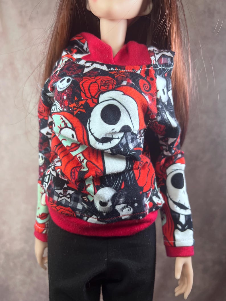 Puede incluir: Una mu&ntilde;eca con una sudadera con capucha estampada en rojo, negro y blanco con capucha y pu&ntilde;os rojos. La sudadera presenta un dise&ntilde;o repetido de calaveras, rosas y otros elementos g&oacute;ticos. La mu&ntilde;eca tambi&eacute;n lleva pantalones negros.